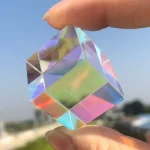 Kristall i kubform med många facetter, transparent med färgade reflexer, hålls mellan två fingrar mot en blå himmel.
