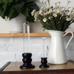 Två laboratorievaser i glas med svarta träbaser, använda som dekorativa objekt, placerade på ett träbord med en bukett vita blommor i en vit keramikkruka.