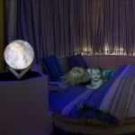 Dekorativ lampa i form av jorden som lyser, placerad på ett bord bredvid en rund säng med två barn som leker och kopplar av i en varm och mysig atmosfär.