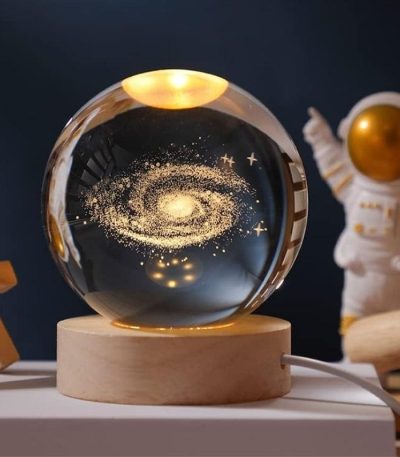 Glaskula som innehåller en miniatyrgalax med stjärnor och kosmisk damm, placerad på en träbas, idealisk för inredning med vetenskapligt eller kosmologiskt tema.