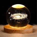 Kristallkula med en representation av en galax i svävande form, belyst och placerad på en träbas, idealisk för vetenskaplig inredning och astronomientusiaster.