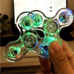 Fidget spinner i färgglad transparent plast, belyst av gröna, blå och röda lampor, hålls av en hand i en inomhusmiljö.