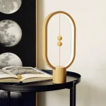 Dekorativ lampa inspirerad av vetenskap, i form av en omloppsbana med gyllene sfärer, placerad på ett svart bord med en öppen bok bredvid.