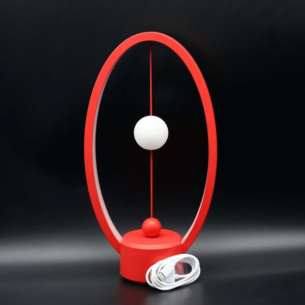 Modern designerklocka för skrivbord med röd oval ram, två hängande vita och röda sfärer, och inkluderad strömkabel, perfekt för vetenskaplig och samtida inredning.