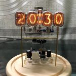 Vintage digital klocka med orange glasrör som visar tiden 20:13:00, på en träbas, i en industriell miljö.