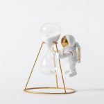 Dekorativ vas i form av sandur med en miniatyrastronaut, innehållande vit sand, på ett gyllene metallstöd, idealisk för vetenskaplig dekoration.