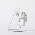Sandur i glas med metallstöd, dekoration i vetenskaplig och rymdinspirerad stil, som representerar en astronaut i vit dräkt som hanterar ett sandur.