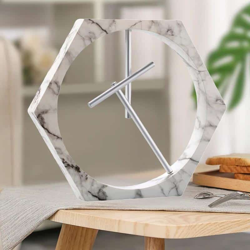 Modern klocka i hexagonform med ram i vit marmor och metallvisare, idealisk för en elegant och samtida inredning.