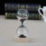 Laboratoriesandur i glas med svart sand, symbol för tidens mätning inom vetenskapen, placerad på en träbas, med en suddig bakgrund av vetenskapsböcker.