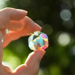 Färgat prismatiskt kristall hålls mellan två fingrar med suddig bakgrund av natur.