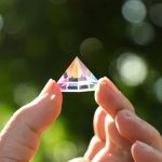 Transparent kristallpyramid hålls mellan två fingrar, reflektion av färgat ljus, suddig bakgrund av grön natur.