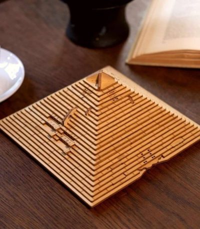 Graverad träpyramidsmodell med vetenskapliga mönster, placerad på ett träbord med en öppen bok och en kaffekopp i närheten.