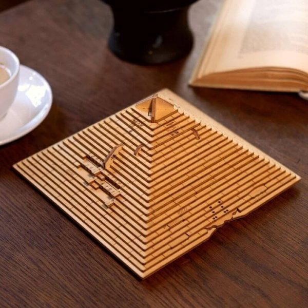 Graverad träpyramidsmodell med vetenskapliga mönster, placerad på ett träbord med en öppen bok och en kaffekopp i närheten.