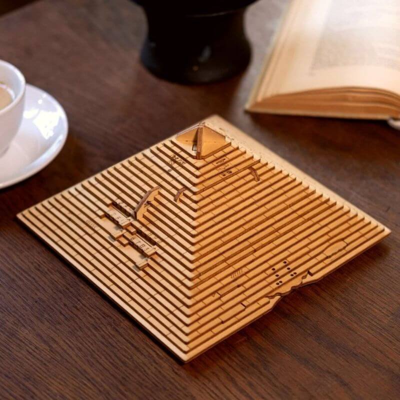 Graverad träpyramidsmodell med vetenskapliga mönster, placerad på ett träbord med en öppen bok och en kaffekopp i närheten.