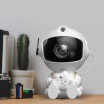 Vit robotkamera med futuristisk design, utrustad med ett vidvinkelobjektiv och en antenn, placerad på ett träbord bredvid en liten kaktusväxt.