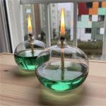 Glasburk som innehåller en grön vätska med en brinnande veke, placerad på en träyta nära ett fönster.