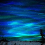 Spektakulär vy av ett norrsken som lyser upp en stjärnklar himmel över ett snöigt landskap, med silhuetter av träd i förgrunden.