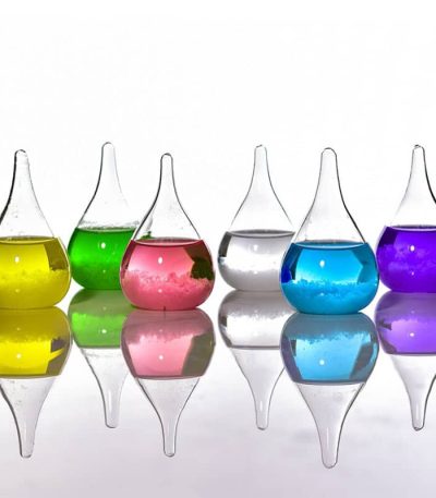 Laboratorievaser i glas med färgade vätskor i gult, grönt, rosa, transparent, blått och lila, placerade på rad med sin spegelbild på en slät yta.