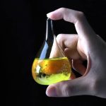 Liten laboratorieflåsa som innehåller en fluorescerande gul vätska, hålls av en hand mot en svart bakgrund, som illustrerar vetenskap och forskning.