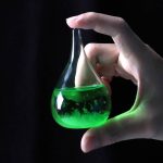 Liten laboratorieflåsa som innehåller en fluorescerande grön vätska, hålls av en hand, mot svart bakgrund.