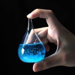 Laboratorievial som innehåller en lysande blå vätska, hålls mellan tumme och pekfinger av en mänsklig hand, mot en svart bakgrund.