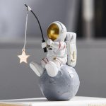 Astronautfigur i vit plast med gyllene hjälm, sittande på en liten grå måne, håller ett fiskespö med en hängande stjärna, dekoration i vetenskaplig och rymdstil.