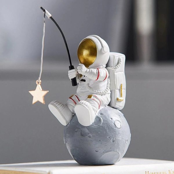 Astronautfigur i vit plast med gyllene hjälm, sittande på en liten grå måne, håller ett fiskespö med en hängande stjärna, dekoration i vetenskaplig och rymdstil.