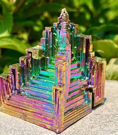 Dekorativ metallbit med iriserande yta, inspirerad av en pyramidformad geometrisk struktur, med färgade reflektioner, placerad på en stenig yta utomhus.
