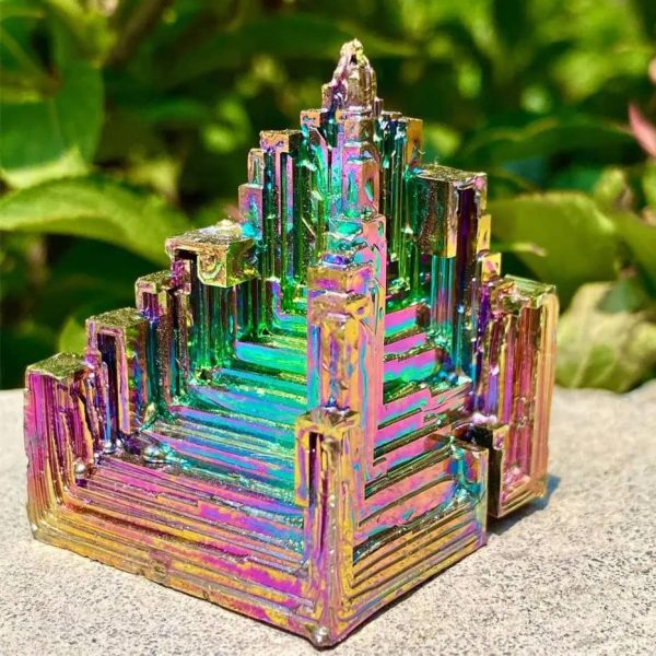 Dekorativ metallbit med iriserande yta, inspirerad av en pyramidformad geometrisk struktur, med färgade reflektioner, placerad på en stenig yta utomhus.