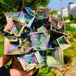 Pyramider av flerfärgade holografiska kristaller, glänsande och geometriska, hållna i handen med en naturlig bakgrund utomhus.