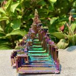 Iriserande kristallpyramid med flerfärgade reflektioner, placerad på en stenyta, omgiven av grön vegetation i bakgrunden.