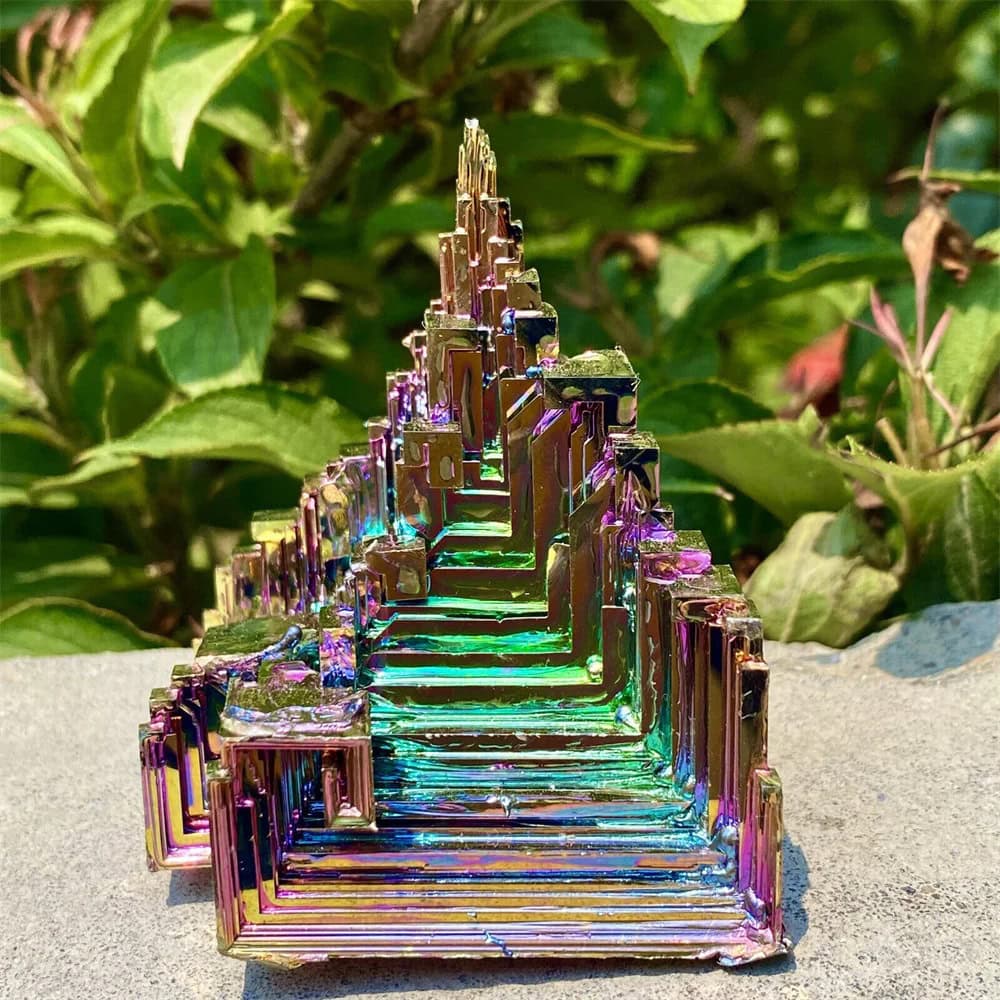 Iriserande kristallpyramid med flerfärgade reflektioner, placerad på en stenyta, omgiven av grön vegetation i bakgrunden.