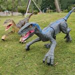 Två gigantiska plysch-T-Rex-dinosaurier för dekoration, placerade på en gräsmatta i en park, med träd i bakgrunden.