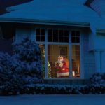 Jultomte klädd i rött, tittar genom det upplysta fönstret i det snöiga huset på natten.