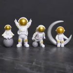 Fyra astronautfigurer i dekoration, med gyllene hjälmar och vita dräkter, tillsammans med en måne i dekoration, perfekta för ett rymd- eller vetenskapligt tema.