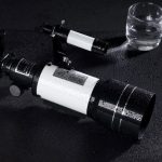 Svart astronomiskt teleskop med bred lins, placerat på en fuktig yta, med ett glas vatten i närheten, som illustrerar vetenskaplig observation och astronomi.