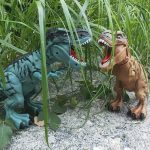 Färgglad T-Rex plyschdinosaurie i en naturlig miljö med gräs och sandig mark.