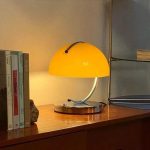 Modern skrivbordslampa med gul skärm, metallbas och böjd arm, som belyser en arbetsyta eller hylla.
