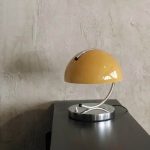 Vintage skrivbordslampa i halvmåneformad gul med metallarm, placerad på en svart yta mot en putsad vägg.