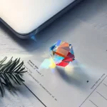 Färgglad prismatisk kristallhänge som projicerar en regnbåge på en vit yta, placerad på ett dokument med en tallgren bredvid.