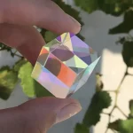 En iriserande kristallkub med flerfärgade reflektioner, hålls mellan två fingrar, med en bakgrund av gröna blad.