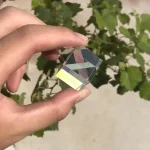 Optisk glassten, kristall eller miniatyr ädelsten hålls mellan fingrarna, med en bakgrund av gröna löv.