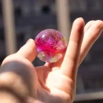 Färgad och genomskinlig glasboll hålls mellan fingrarna, med en suddig bakgrund av en stadsscen. Perfekt för vetenskaplig dekoration eller som samlarobjekt.