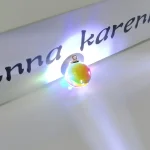 Färgad glasberlock med inbyggd LED-lampa, placerad på en vit yta, med en delvis textbakgrund "anna karen".