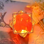 Hänge i kubform med upplysta orange stenar, inramat av en gyllene metallstruktur, placerat på en träyta med en suddig bakgrund av blommor och natur.