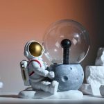 Astronautfigur i vetenskapsdekoration, sittande bredvid en glödlampa i en minimalistisk miljö.