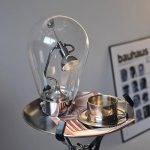 Dekorativ lampa i form av en glödlampa med synligt filament, placerad på en bricka med en metallmugg.