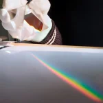 Tång som håller ett optiskt prisma som skapar en regnbåge av ljus på en slät yta, som illustrerar ljusets spridning.