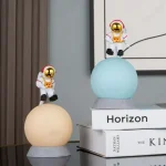 Två plastastronautfigurer med gyllene hjälmar, placerade på lysande sfärer, dekorerar en modern inredning med böcker i bakgrunden.