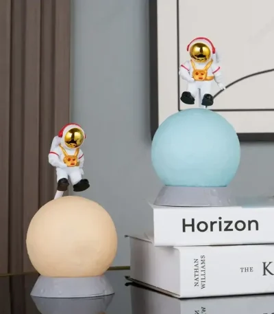 Två plastastronautfigurer med gyllene hjälmar, placerade på lysande sfärer, dekorerar en modern inredning med böcker i bakgrunden.